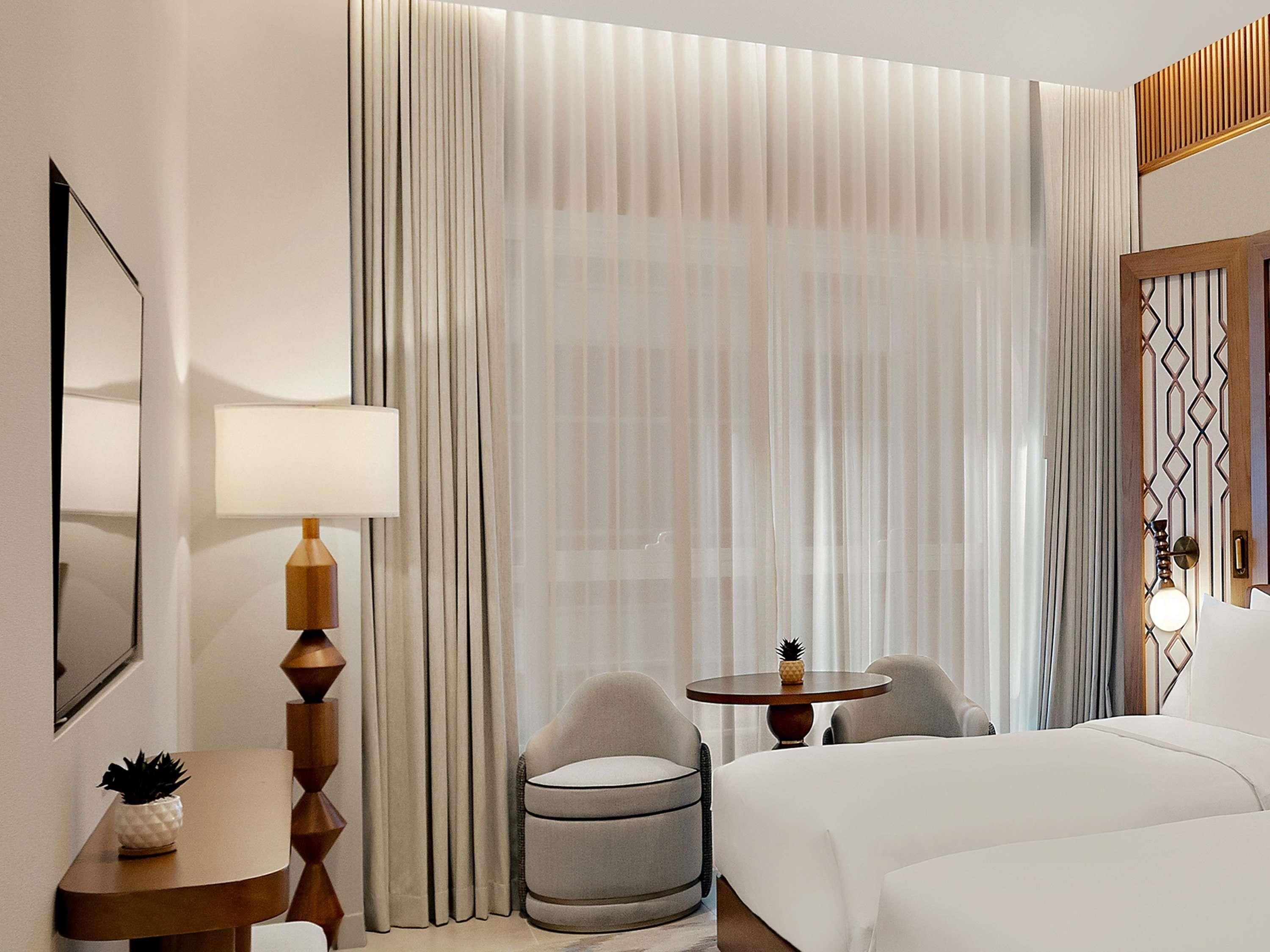 Une chambre d’hôtel avec un lit, une table ronde et deux chaises, une petite table d’appoint avec une lampe, et une grande fenêtre avec des rideaux.