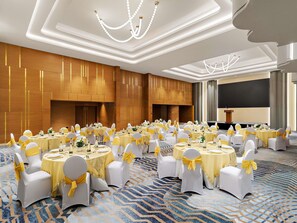Une salle de banquet avec des tables rondes, des chaises blanches ornées de rubans jaunes et un grand écran.