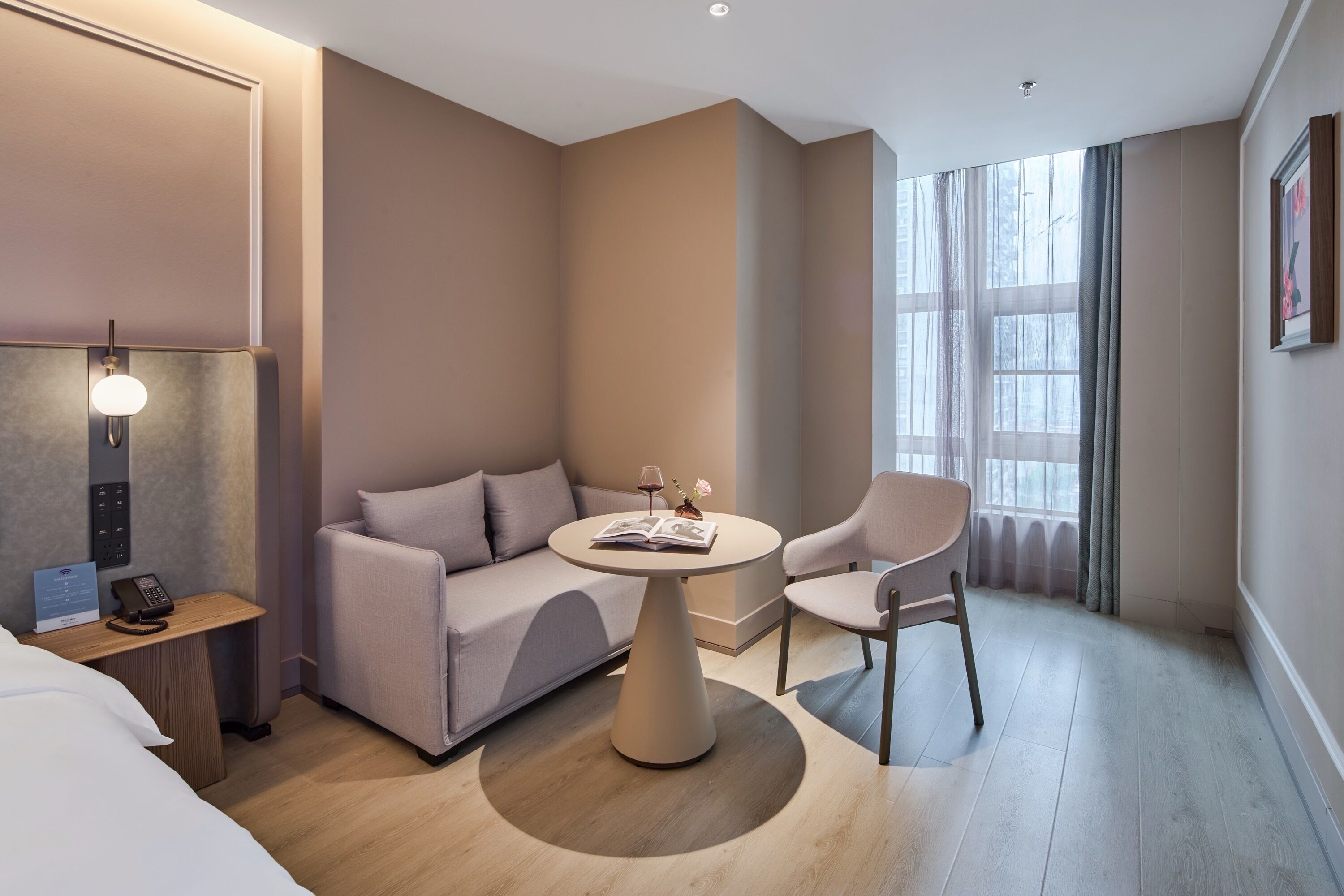 A modern hotel room with a sofa, a round table, a chair, a bedside table with a lamp, and a window with curtains.