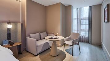 A modern hotel room with a sofa, a round table, a chair, a bedside table with a lamp, and a window with curtains.