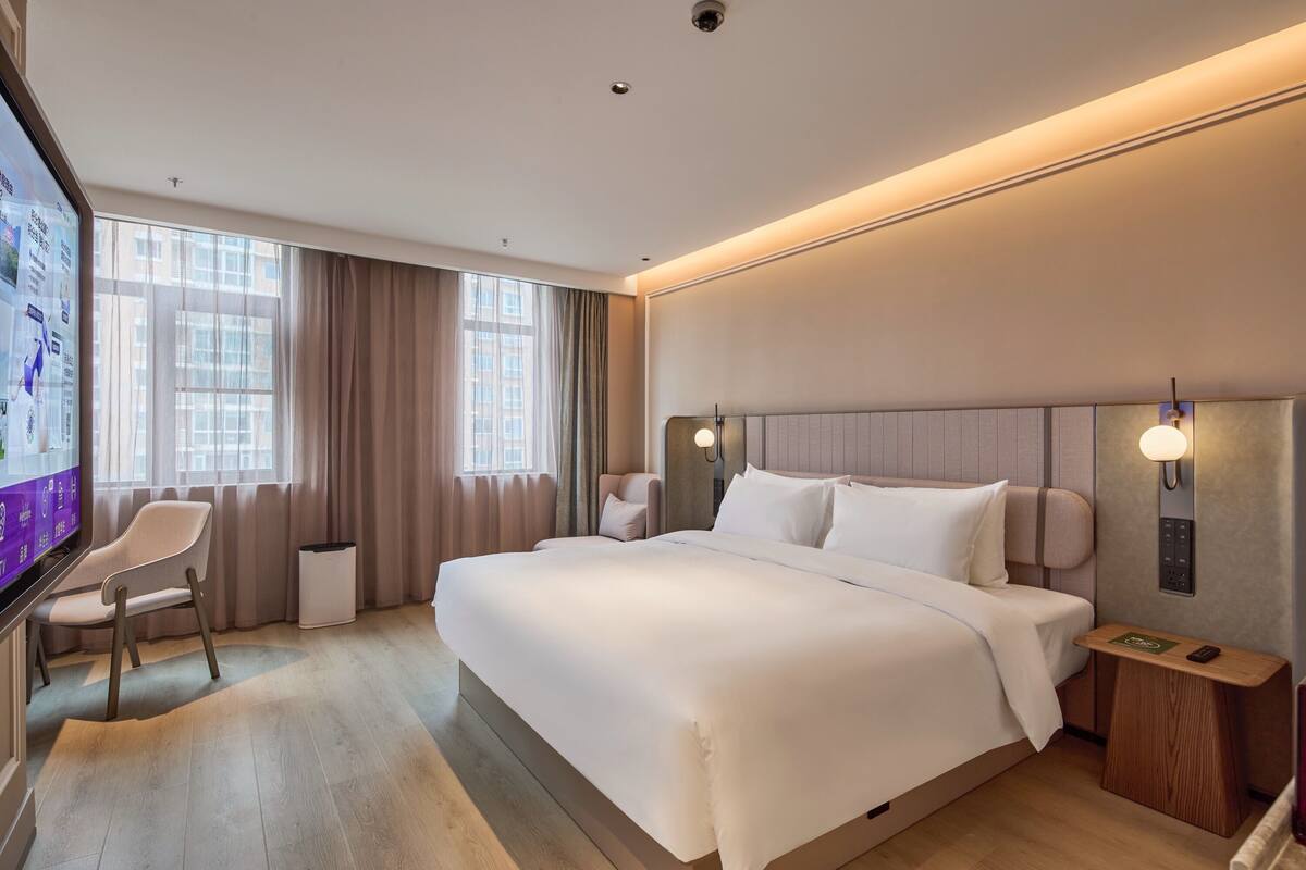 A modern hotel room with a large bed, a desk with a chair, a TV, and a view of the city through large windows.