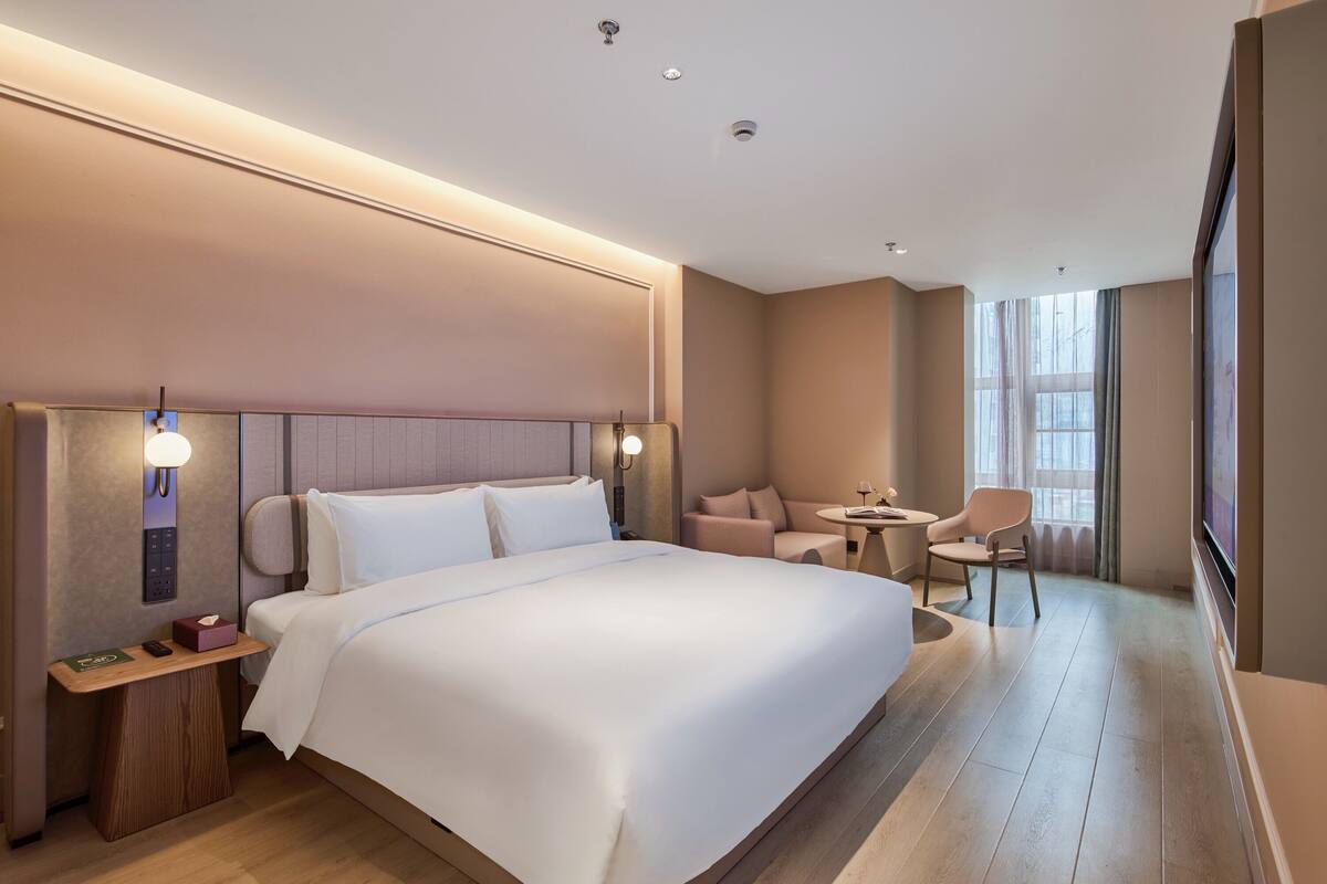 A modern hotel room with a large bed, bedside tables, a seating area with a chair and table, and a large window.