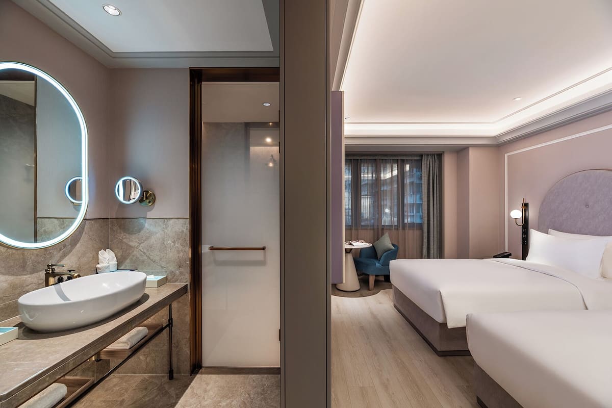 A hotel room with a large bed, a bedside table, a bathroom with a sink and mirror, and a view of the city through the window.