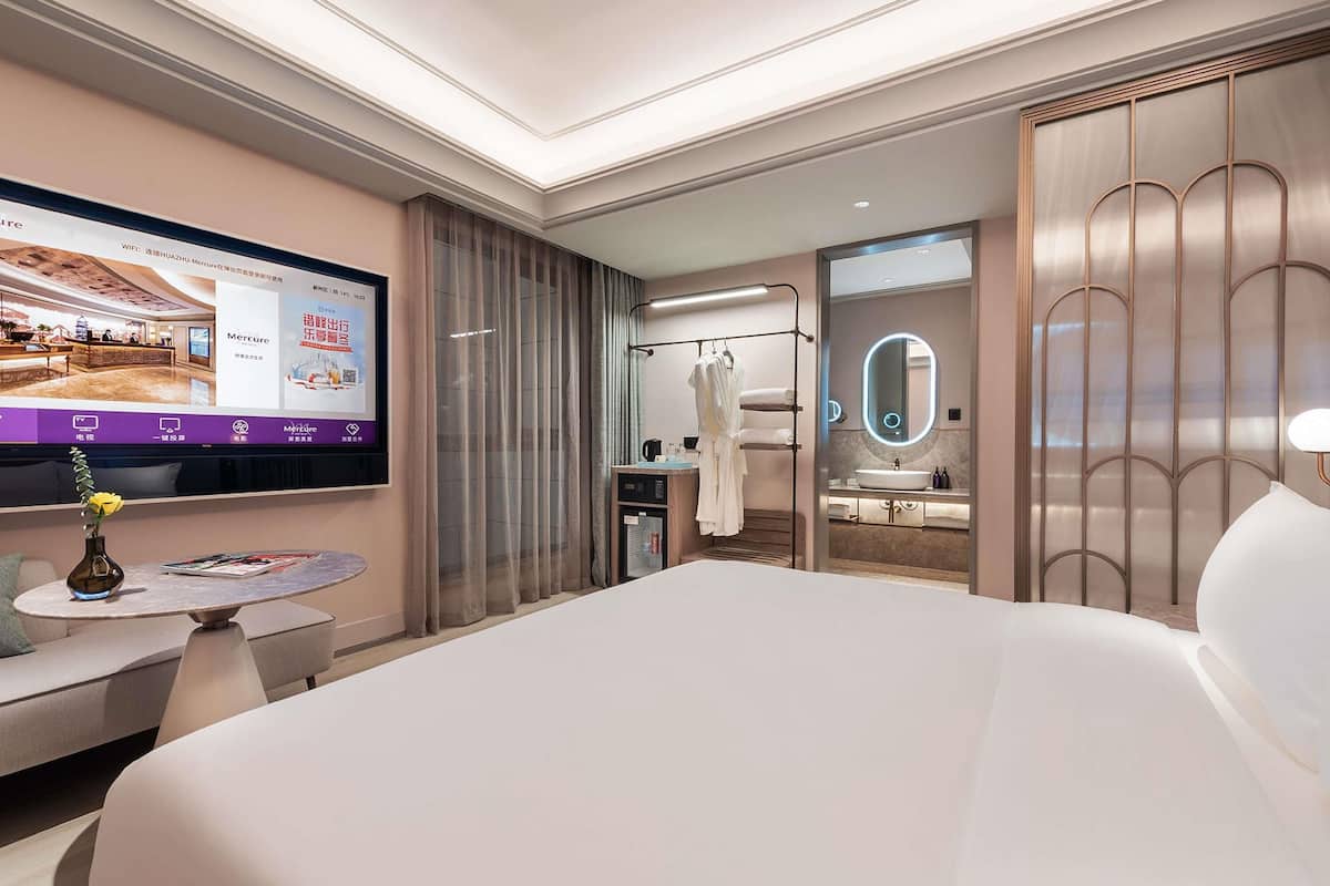 A modern hotel room with a large bed, a TV mounted on the wall, a bathroom with a vanity and mirror, and a seating area with a small table and a vase.