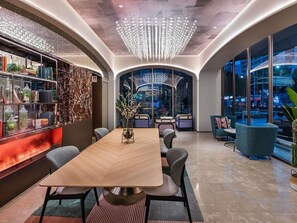 A modern conference room with a wooden table, chairs, a chandelier, a bookshelf, and large windows with a city view.