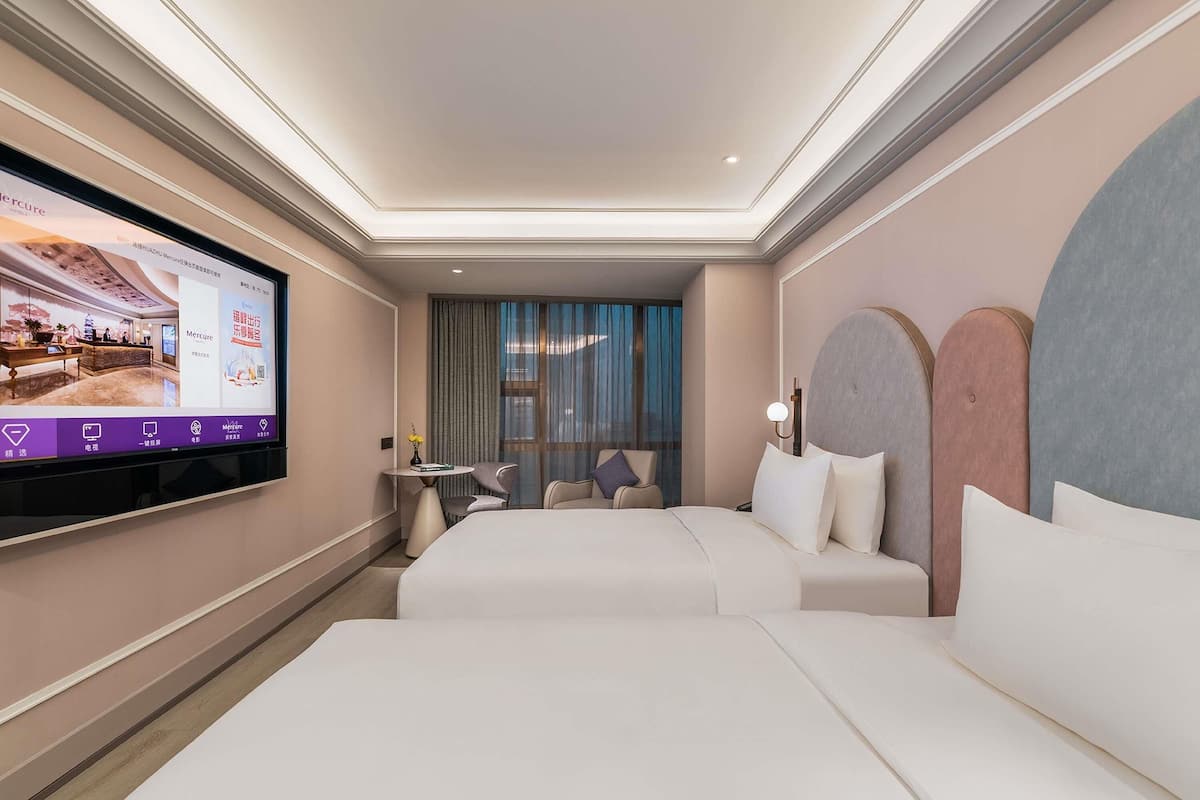 A hotel room with a large flat-screen TV, two beds, a small table with a vase, and a view of the city through large windows.