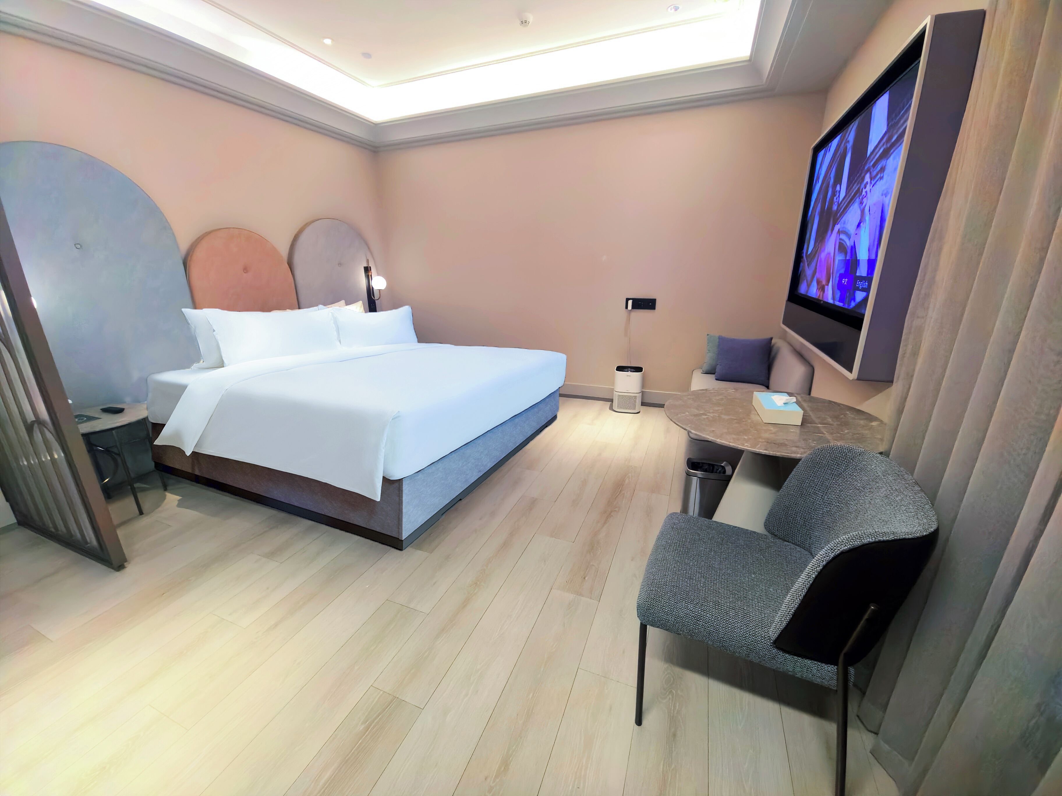 A modern hotel room with a bed, a desk, a chair, a TV, and a trash bin.