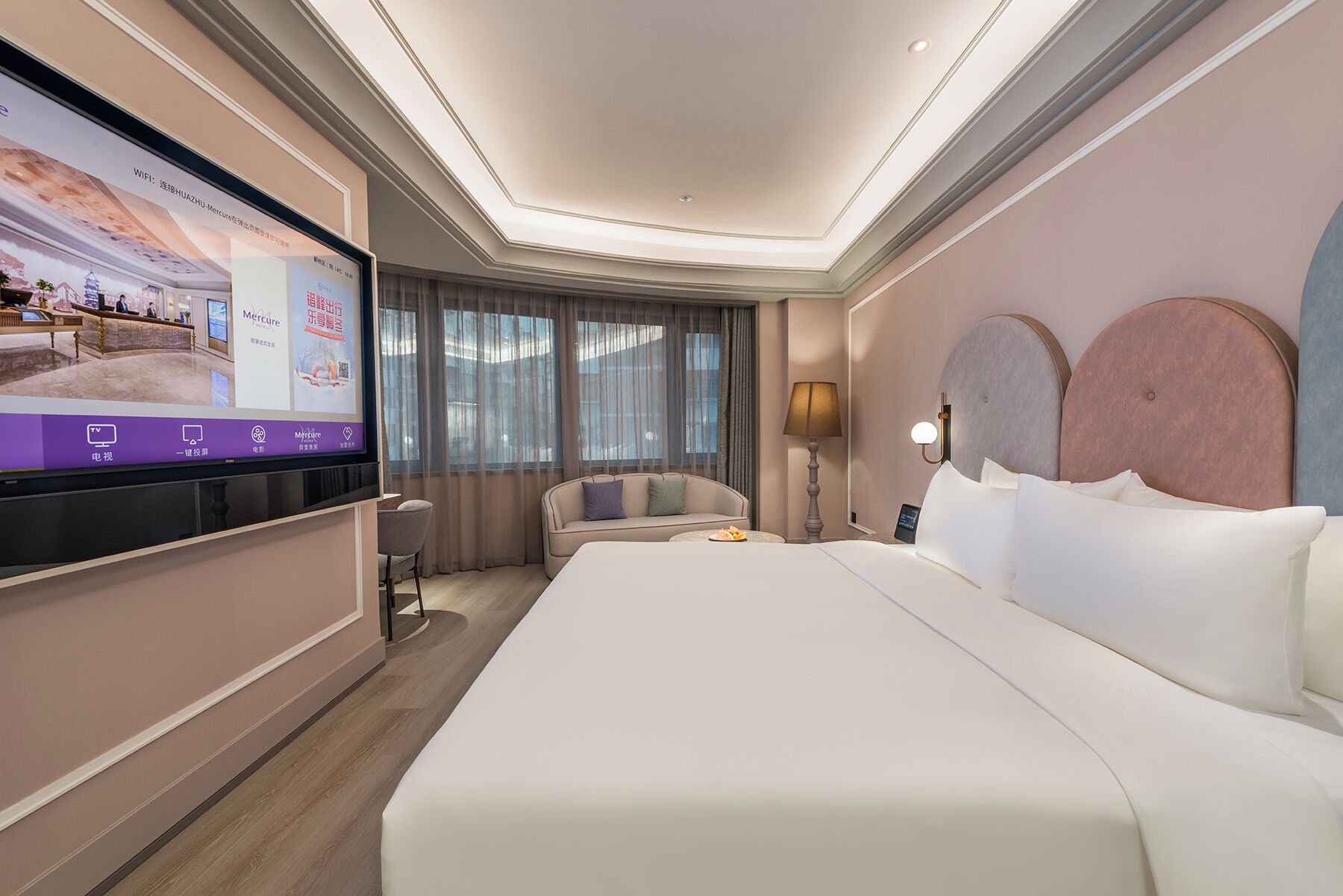 Une chambre d’hôtel moderne avec un grand lit, un canapé, un téléviseur fixé au mur et une fenêtre avec des rideaux.