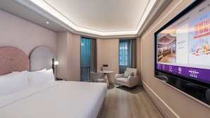 Ein Hotelzimmer mit einem großen Bett, einem Schreibtisch, zwei Stühlen, einem Fernseher der Hotelinformationen anzeigt und einer Aussicht nach draußen durch das Fenster.