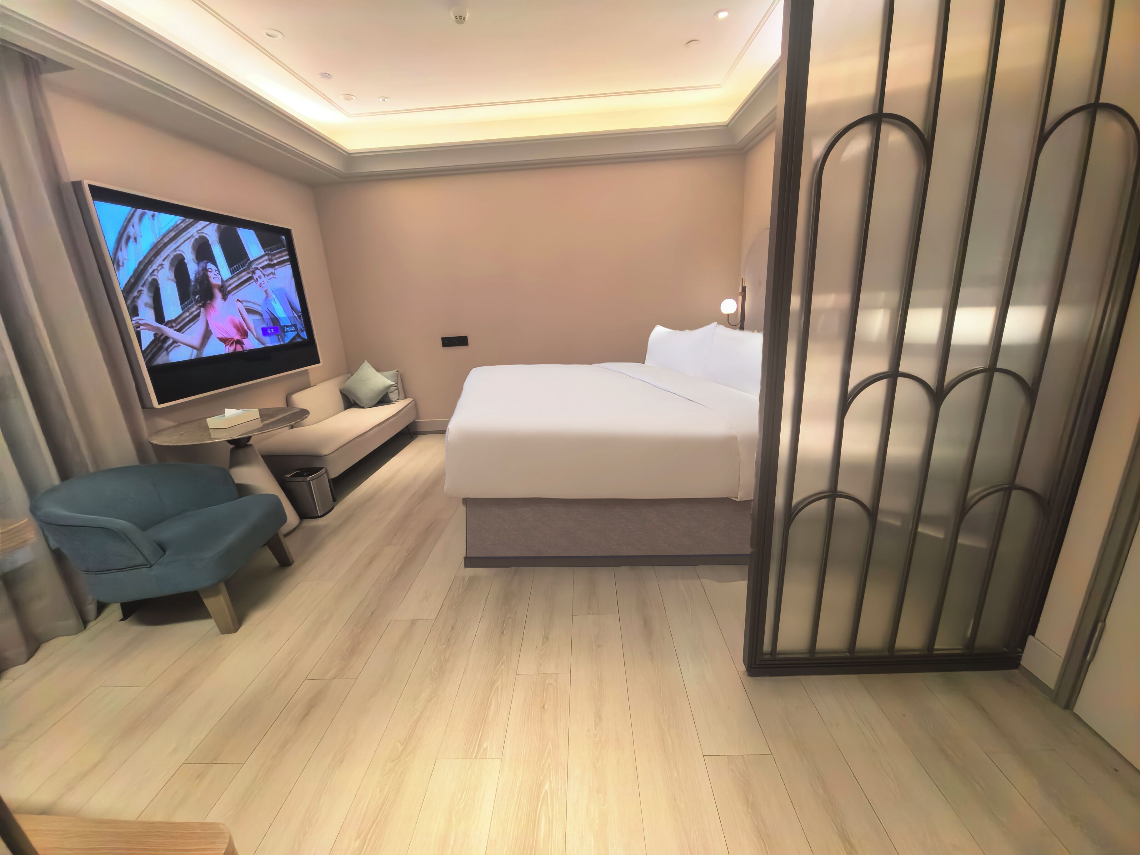 A modern hotel room with a bed, a TV, a seating area, and a decorative partition.