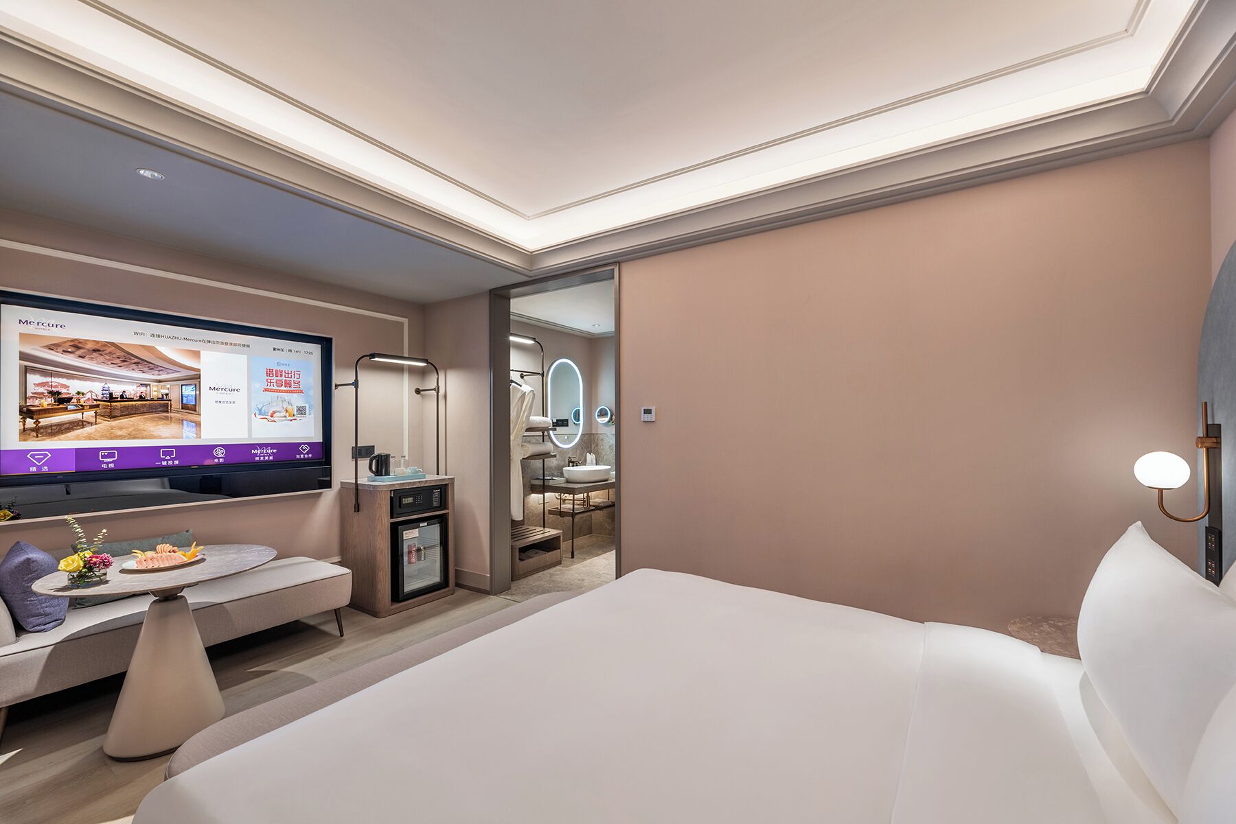 Une chambre d’hôtel moderne avec un grand lit, un bureau avec un ordinateur, une télévision et une salle de bain visible à travers une porte ouverte.