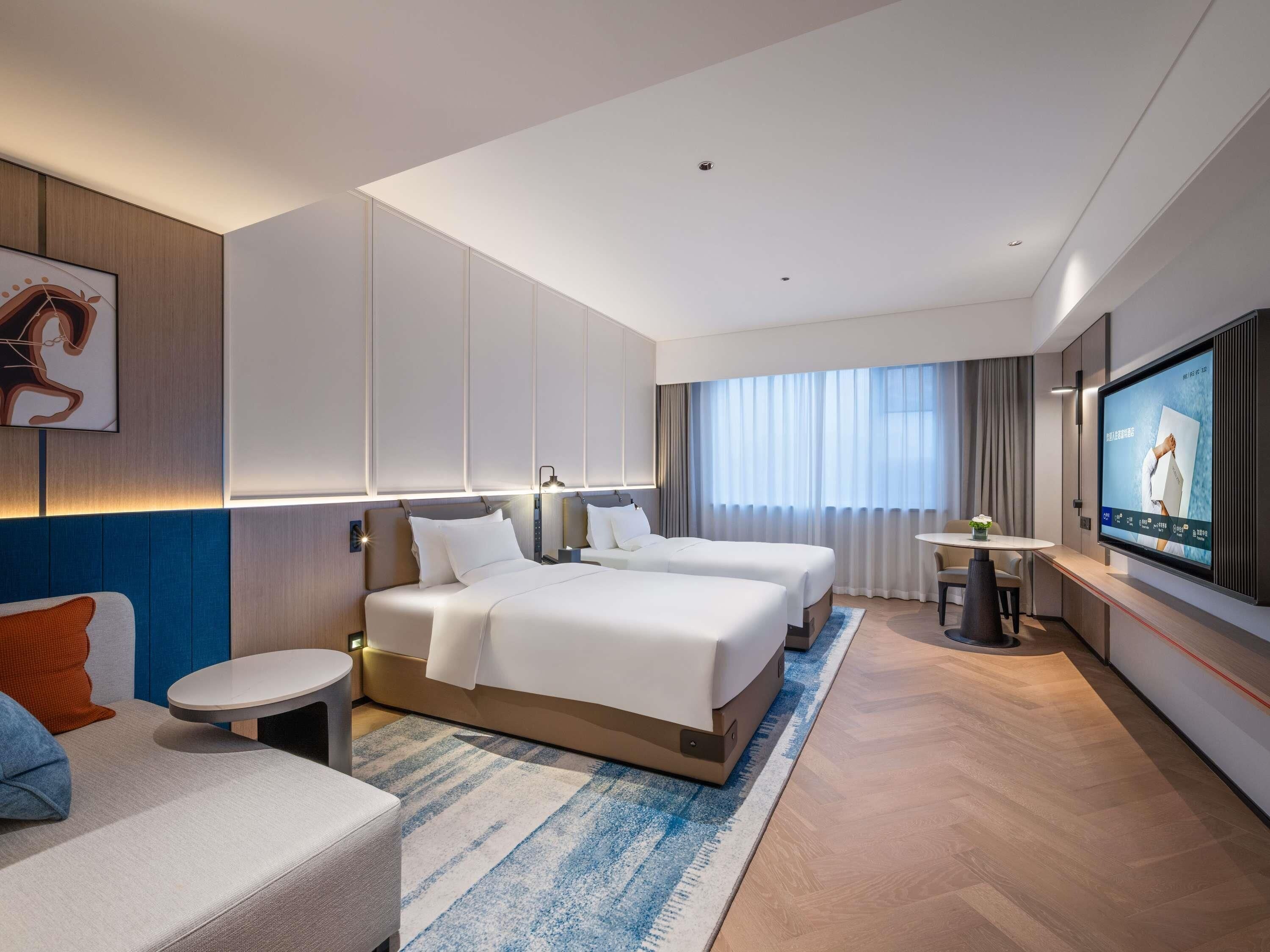 A modern hotel room with two beds, a desk, a television, and a large window with curtains.