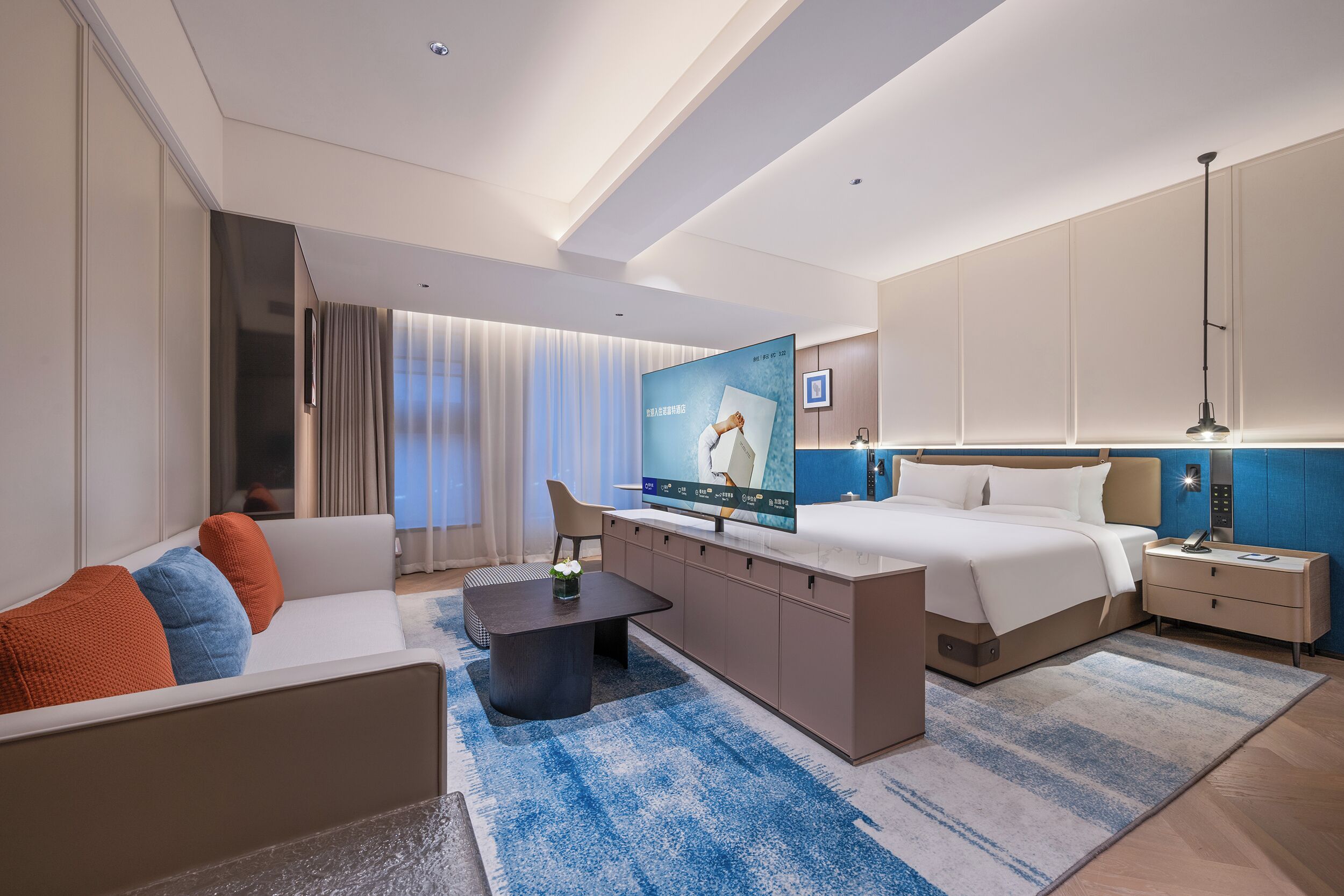 A modern hotel room with a large bed, a sofa, a coffee table, a TV, and a desk with a lamp.