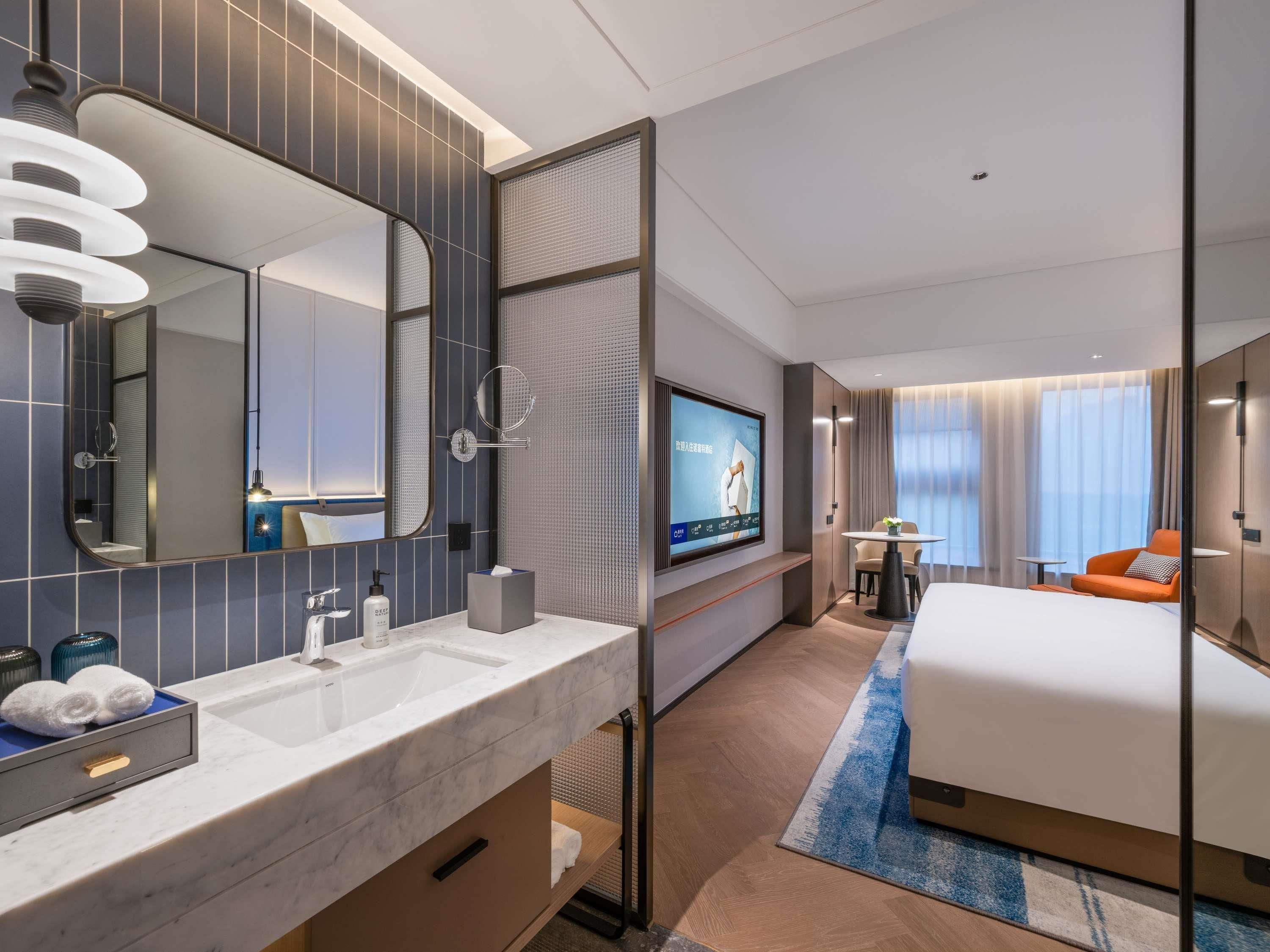 Una camera d'albergo con un letto grande, una TV a muro, un bagno con specchio, lavandino e porta asciugamani.