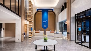 A modern hotel lobby with a reception desk, a blue vertical sign, and a round table with a plant. - Novotel Hangzhou Yuhang (Hangzhou)