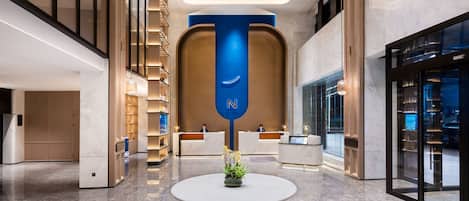 Un moderno vestíbulo de hotel con una recepción, un letrero vertical azul y una mesa redonda con una planta.