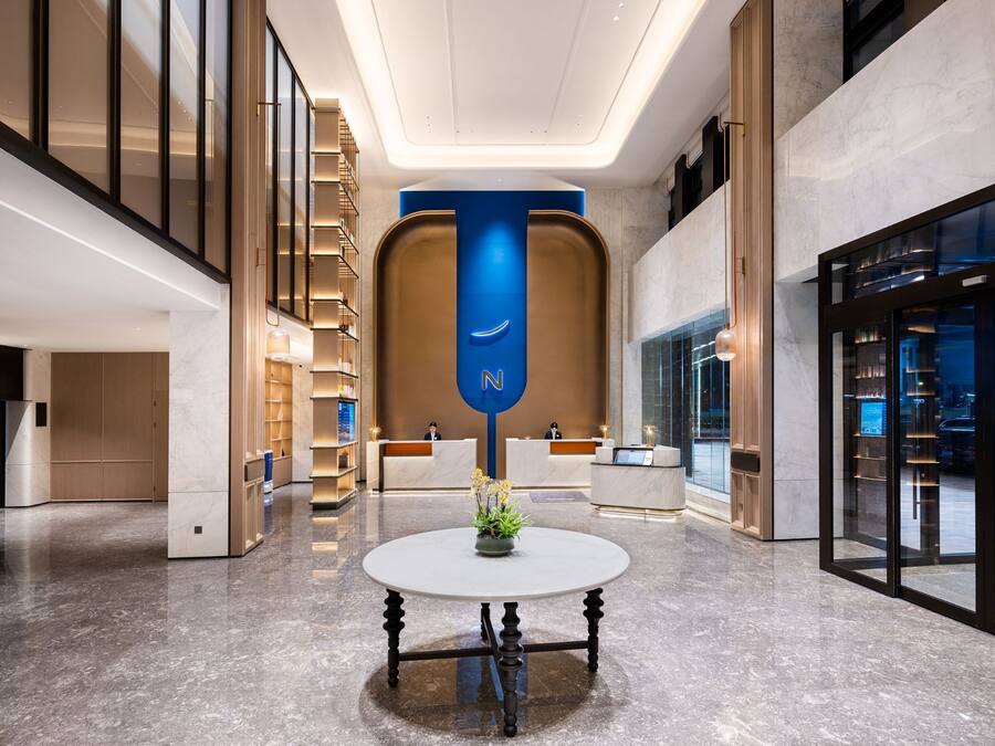 A modern hotel lobby with a reception desk, a blue vertical sign, and a round table with a plant.