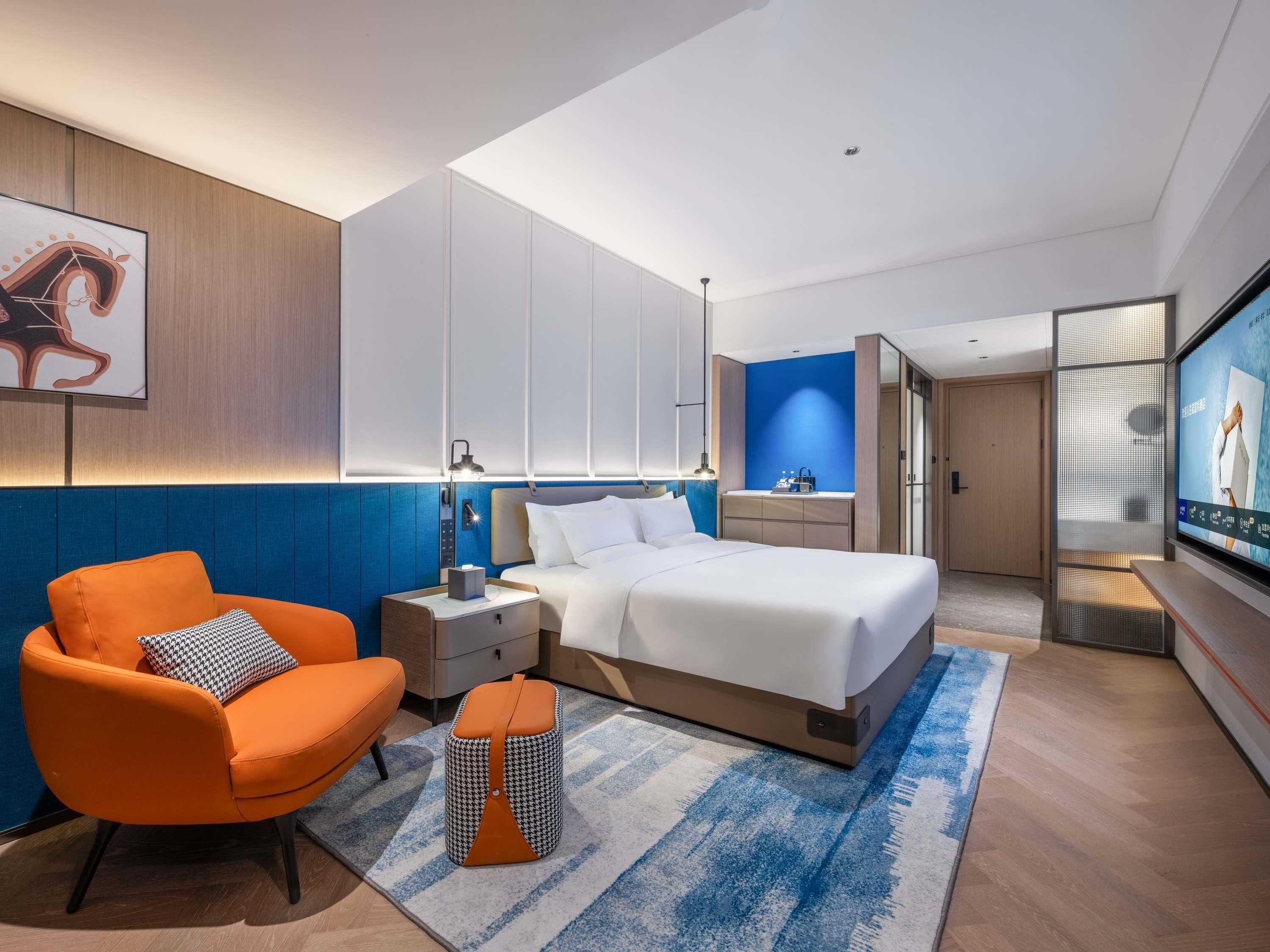 Une chambre d’hôtel moderne équipée d’un lit, d’une table de chevet, d’une chaise, d’un petit pouf et d’un téléviseur.