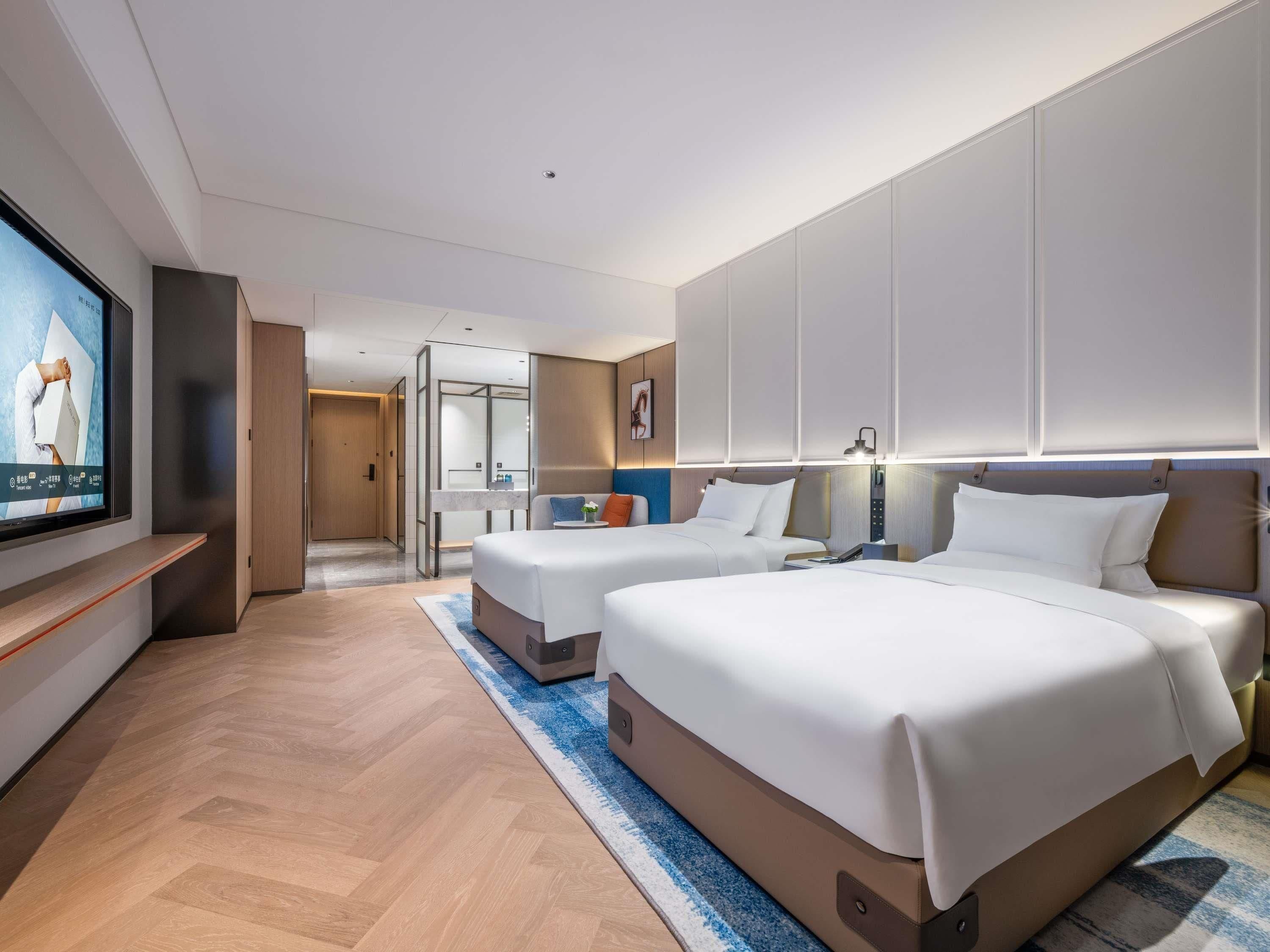 Ein Hotelzimmer mit zwei Betten, einem Fernseher, einem Schreibtisch und einem durch eine Glaswand abgetrennten Badezimmer.