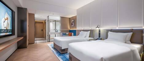 A hotel room with two beds, a TV, a desk, and a bathroom visible through a glass partition.