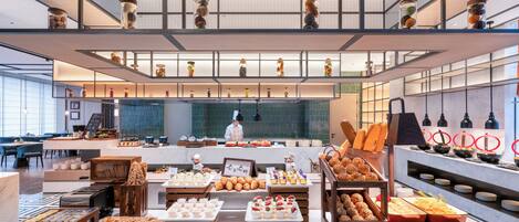 A modern restaurant with a chef preparing food, various food displays, and a high ceiling with hanging lights.