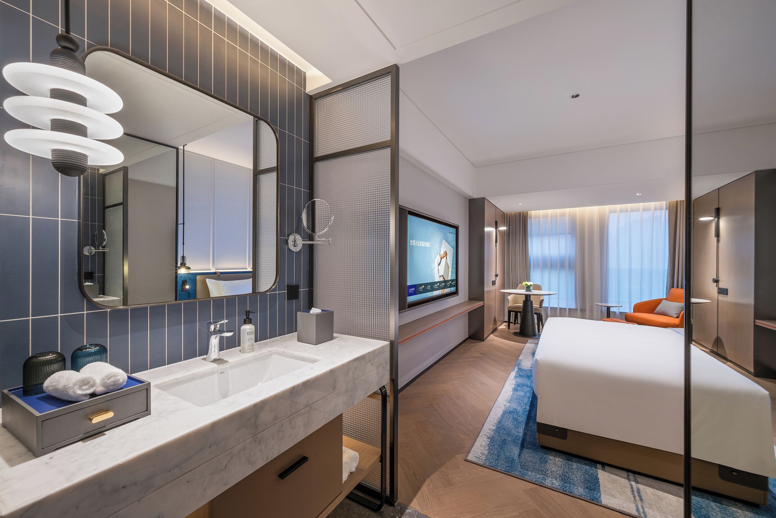 A modern hotel room with a large bed, a TV, a bathroom with a mirror, and a seating area with a table and chairs.