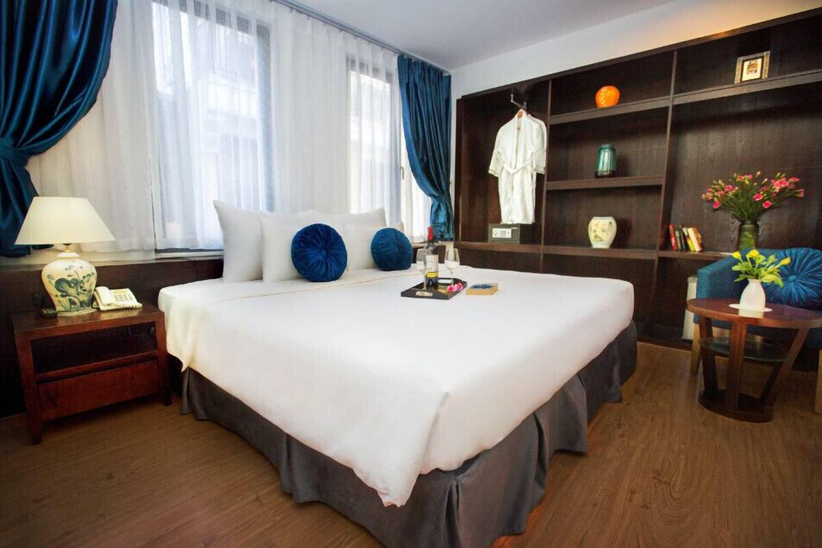 Hotellihuone, jossa on suuri sänky, yöpöytä lampun kanssa, vaatekaappi ja pieni pöytä, jossa on maljakko täynnä kukkia.