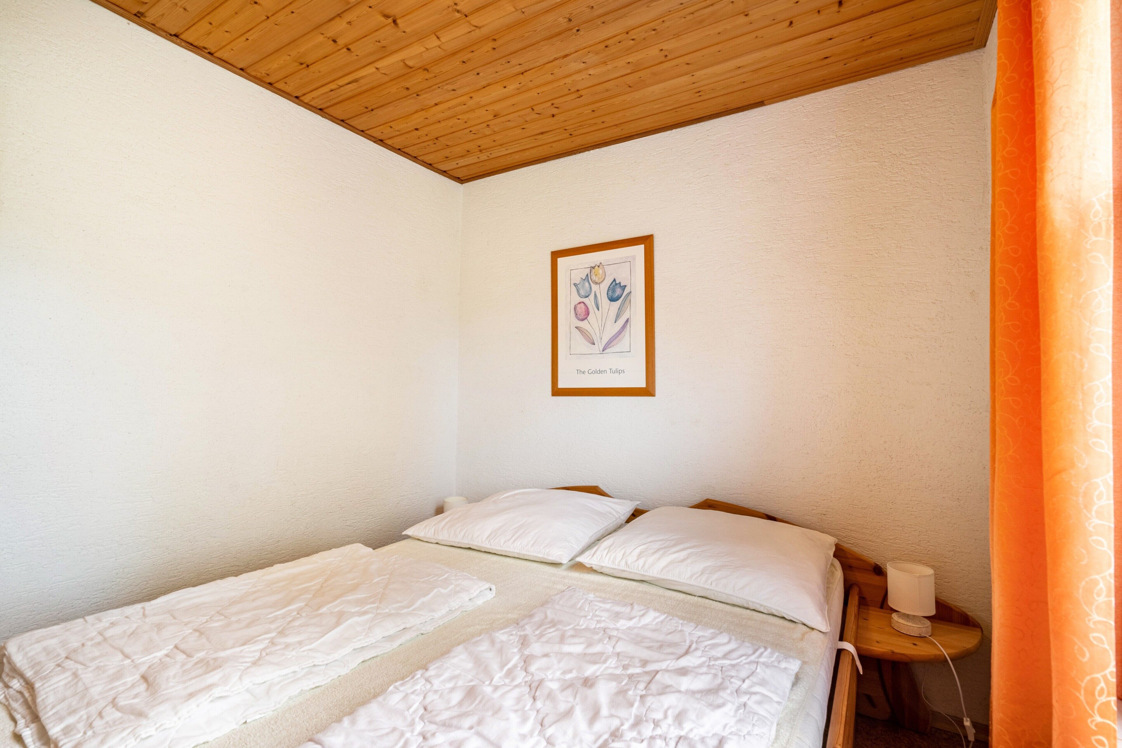 A room with a wooden ceiling, a bed with white bedding, a framed picture on the wall, and a nightstand with a lamp.