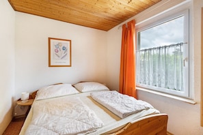 Une chambre avec un plafond en bois, une grande fenêtre ornée de rideaux orange, un tableau encadré au mur et un lit avec une literie blanche.