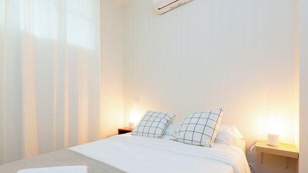 A neatly made bed with white linens and checkered pillows, a bedside table with a lamp, and a wall-mounted air conditioner. - Apartment Mirage Villa Cana (Cannes)