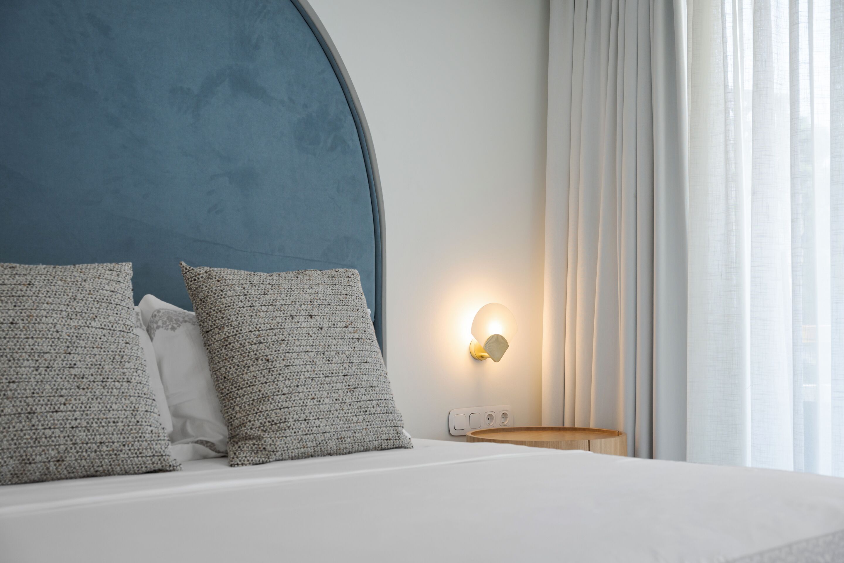 Una cama bien hecha con tablero de cabecera azul, dos almohadas con textura, una mesita de noche con lámpara y una ventana con cortinas traslúcidas.