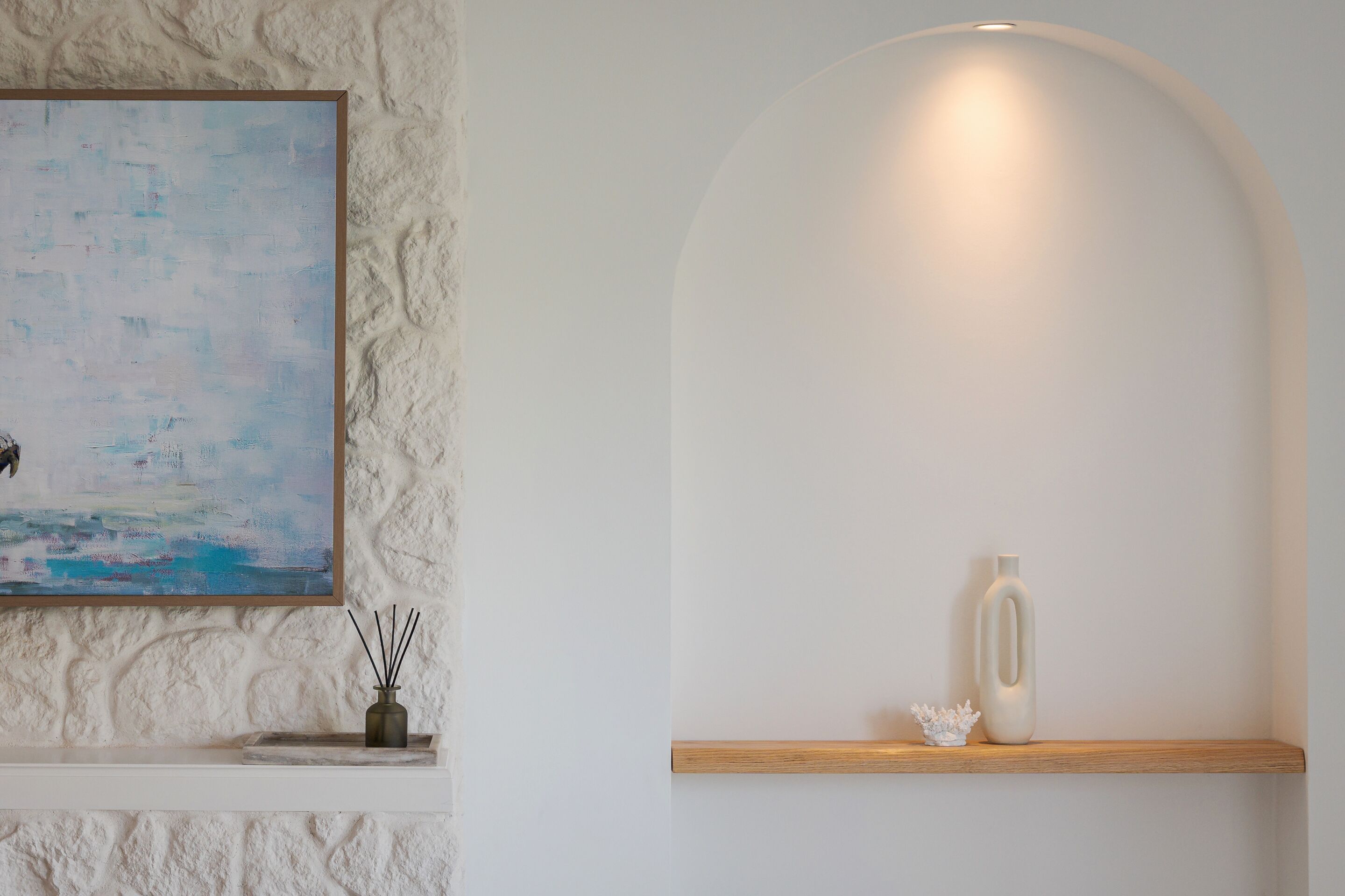 Ein Zimmer mit strukturierter Wand, einem Gemälde, einem Regal mit Vase und Kerze sowie einer gewölbten Wand mit einer Leuchte.