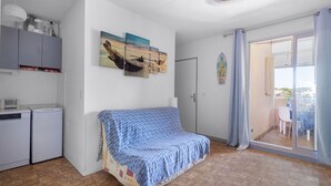 Una habitación con sofá, kitchenette, puerta corrediza de vidrio a un balcón y una pared con un cuadro.
