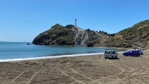 A rocky beach with a lighthouse on a hill, a van, and a blue car parked on the sand. - Waiwhare On The Beach (beachfront property with direct beach access) (Castlepoint)