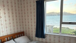 A bedroom with a bed, floral wallpaper, a window with a view of a body of water and mountains, and a wooden headboard. - Waiwhare On The Beach (beachfront property with direct beach access) (Castlepoint)