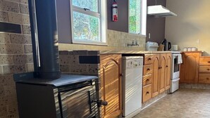 A kitchen with a wood-burning stove, wooden cabinets, a dishwasher, and a window overlooking greenery. - Waiwhare On The Beach (beachfront property with direct beach access) (Castlepoint)