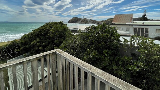 A coastal view with a wooden deck overlooking the sea, a building with a satellite dish, and a mountainous backdrop. - Waiwhare On The Beach (beachfront property with direct beach access) (Castlepoint)