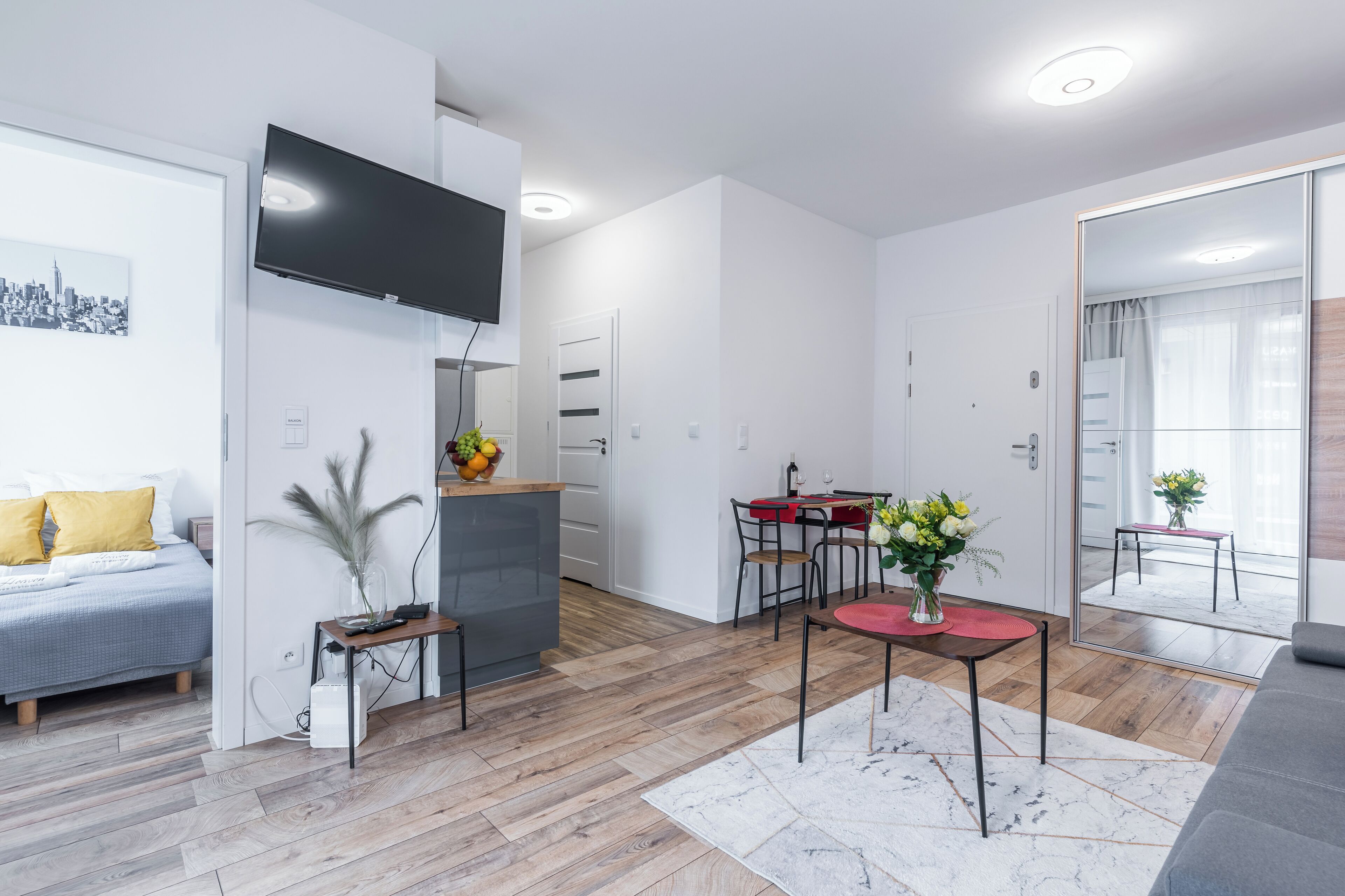 Una sala de estar moderna con un televisor de pantalla plana, una zona de comedor con una mesa roja y un espejo grande que refleja la habitación.