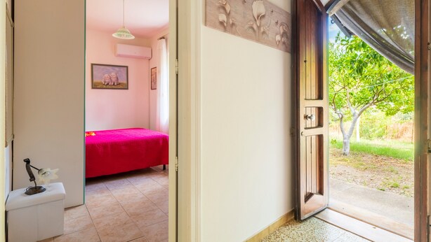 Puerta abierta que da a un dormitorio con colcha roja, una mesita blanca con una flor y un cuadro colgado en la pared.
