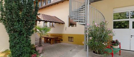 A residential building with a spiral staircase, a patio with a table and bench, and a variety of potted plants.