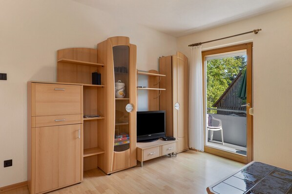 Une chambre avec une armoire en bois, une télévision et une porte-fenêtre coulissante donnant sur un balcon équipé d’une chaise.