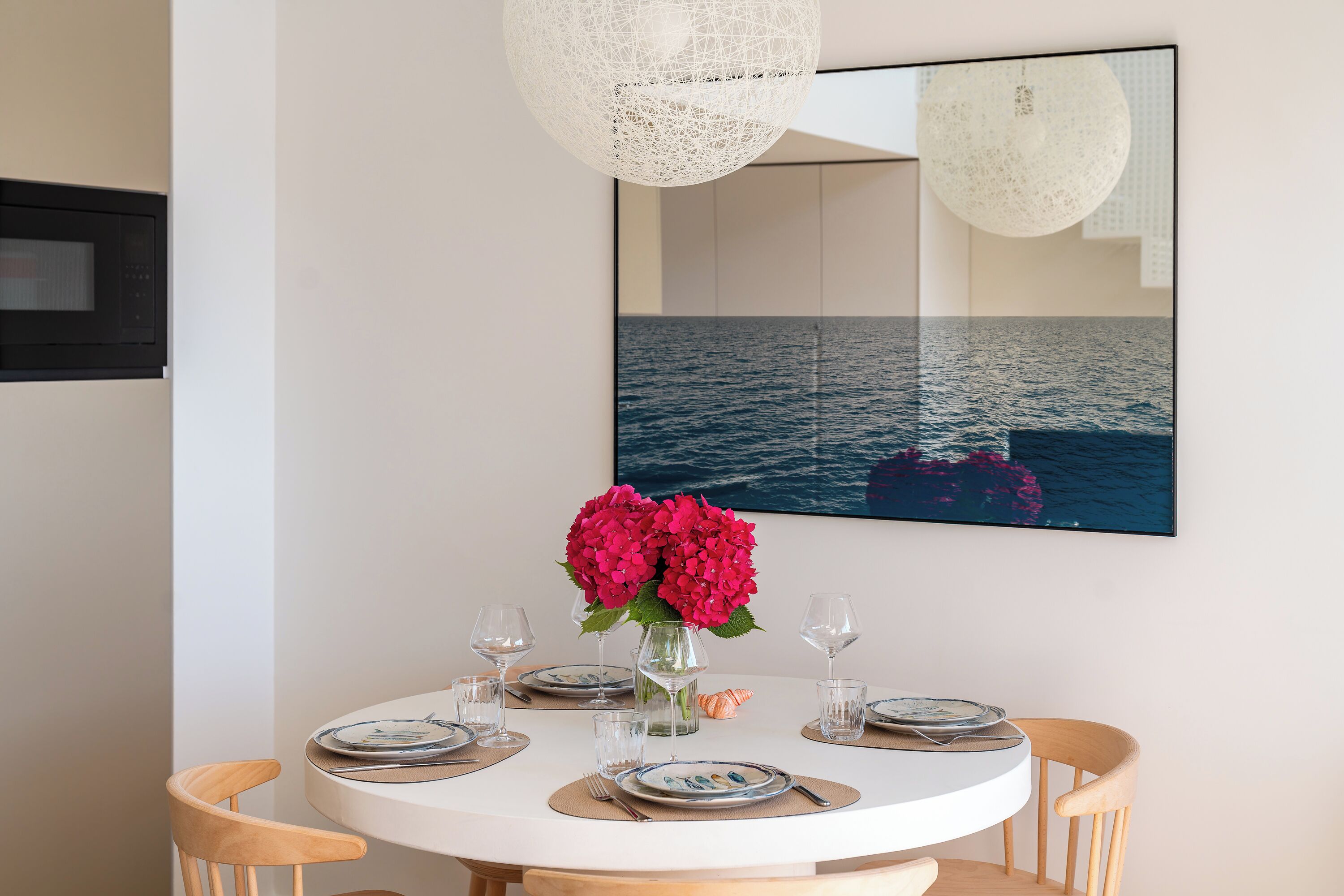 Un coin repas avec une table ronde dressée pour quatre personnes, un vase de fleurs et un grand tableau encadré représentant la mer, accroché au mur.