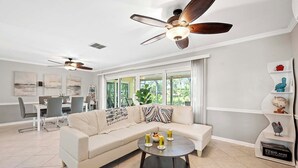 Ein modernes Wohnzimmer mit einem beigefarbenen Ecksofa, einem runden Couchtisch mit Getränken, einem Essbereich mit weißem Tisch und Stühlen sowie einem Deckenventilator.