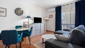 Un salon avec un canapé, une table à manger et des chaises, un téléviseur, une horloge et une fenêtre avec des rideaux.