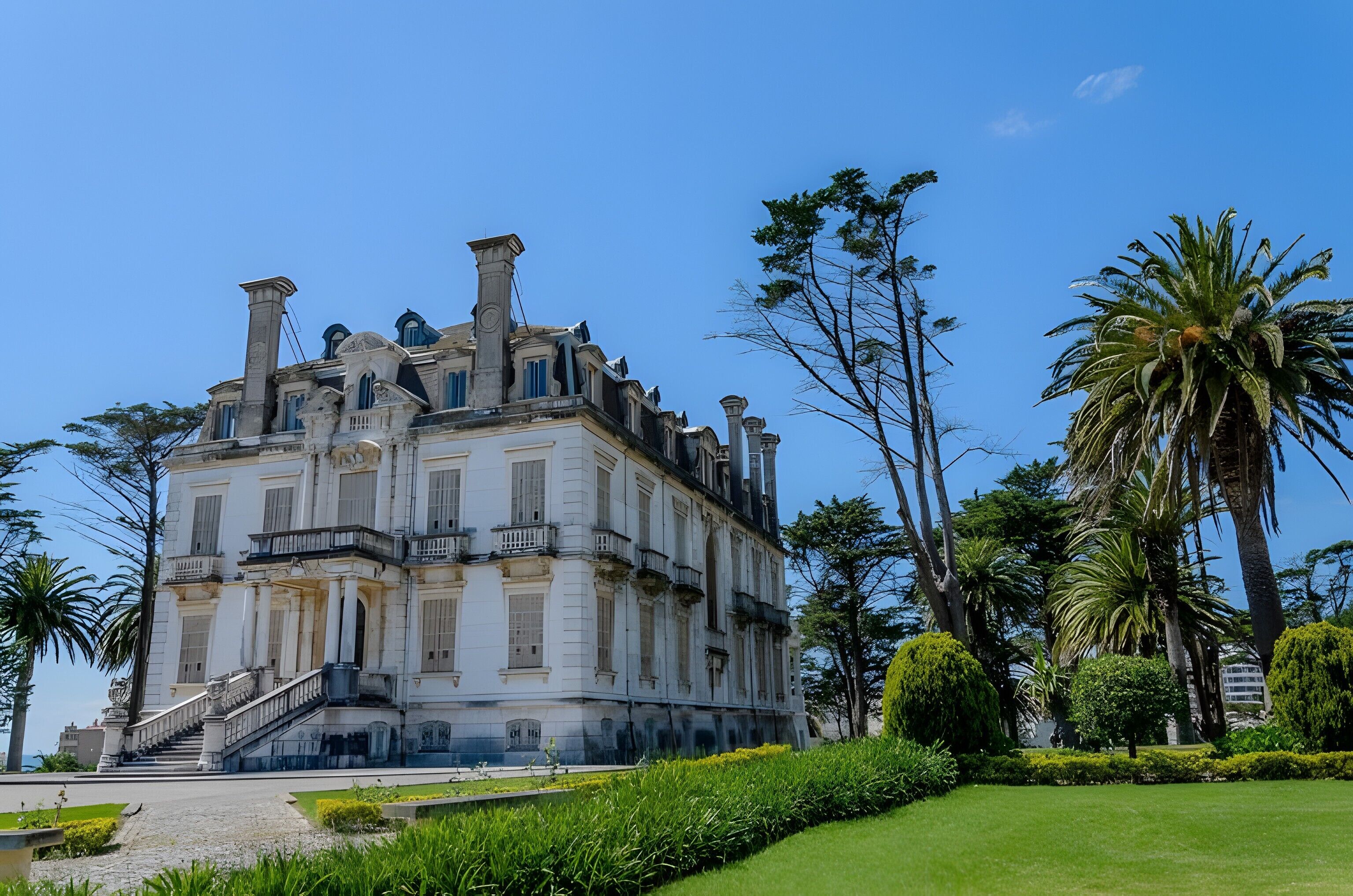 Um edifício grande e histórico com uma escadaria imponente, rodeado por jardins bem cuidados e palmeiras.