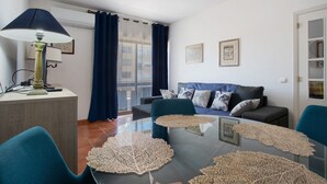 Un salon moderne avec une table basse en verre, des fauteuils bleus, un canapé agrémenté de coussins à motifs et une lampe posée sur une table d’appoint en bois.