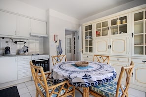Une cuisine avec des meubles blancs, une table à manger recouverte d’une nappe à motifs, des chaises en osier et un panier posé sur la table.