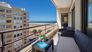 Un balcon avec vue sur la mer, des meubles en osier, une table avec un livre et des verres, et une plante en pot.