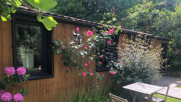 A wooden cabin with a window, a table with chairs, and pink flowers growing on the wall.