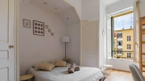 Ein Schlafzimmer mit einem Bett, einem Nachttisch, einer Lampe und Blick auf Gebäude durch ein Fenster.